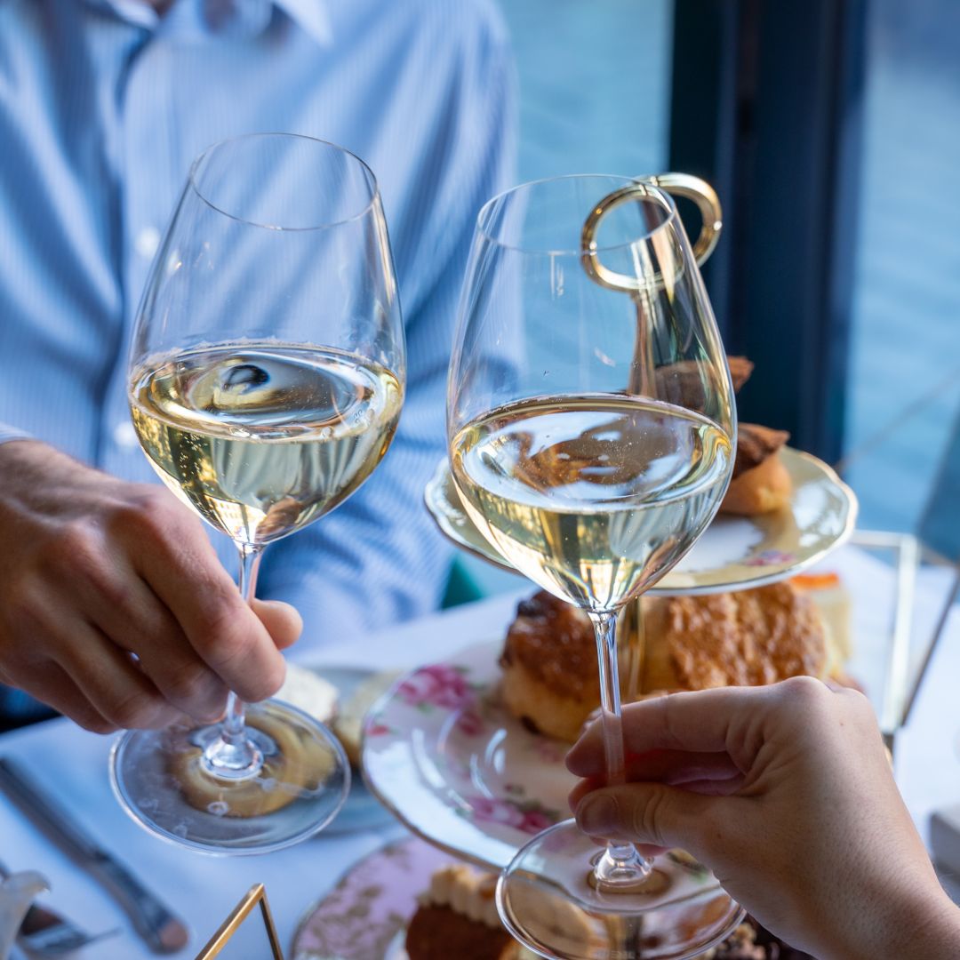 Two people clinking glasses of white wine over a table with pastries on a floral plate.